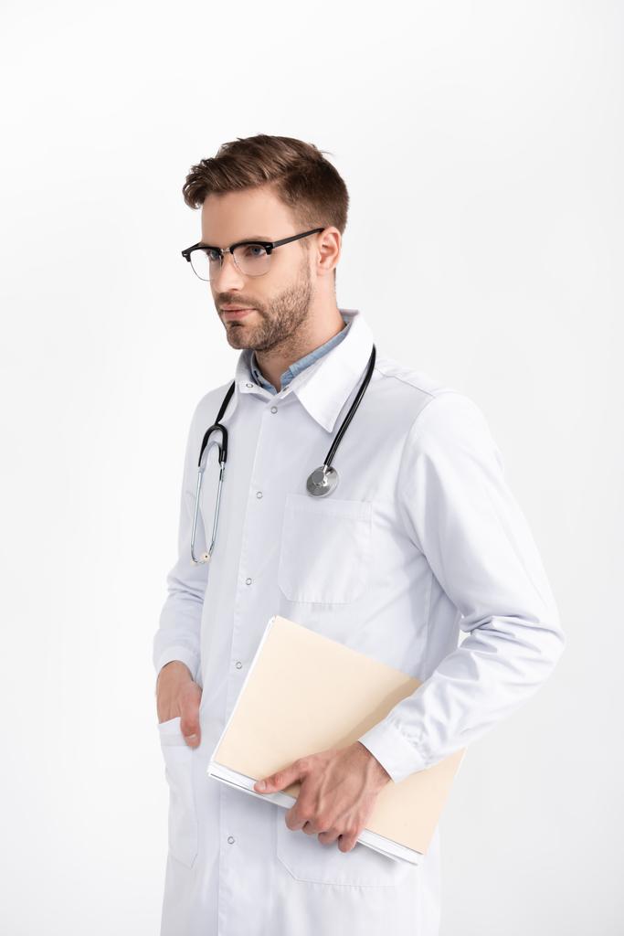 Médico con estetoscopio con la mano en el bolsillo mirando hacia otro lado, mientras sostiene la carpeta aislada en blanco - Foto, Imagen