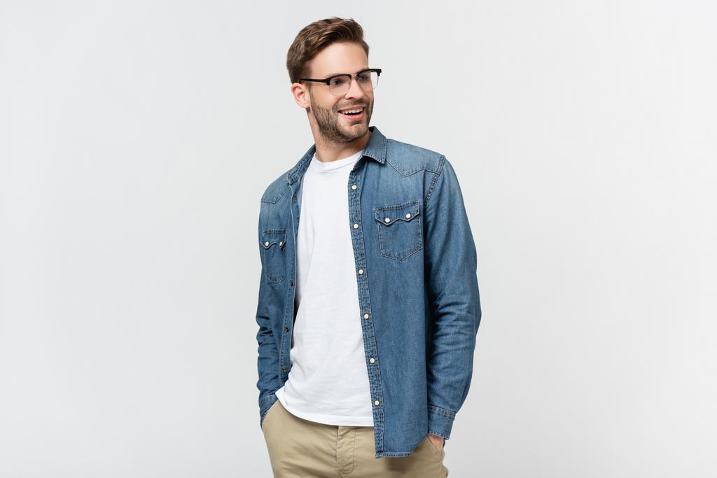 Hombre sonriente en gafas con las manos en los bolsillos de los pantalones mirando hacia otro lado aislado en gris - Foto, Imagen