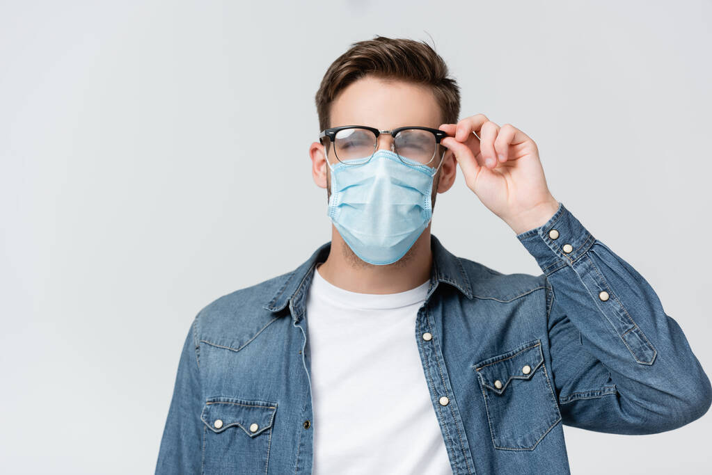 Joven hombre con máscara médica tocando gafas empañadas aisladas en gris - Foto, Imagen