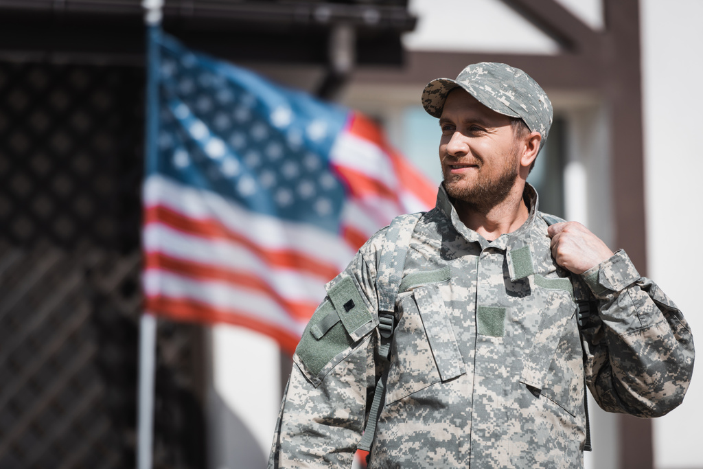 Uśmiechnięty weteran wojskowy w kamuflażu odwracając wzrok zamazaną amerykańską flagą na tle - Zdjęcie, obraz