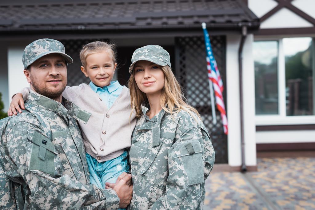 Усміхнений батько і мати у військовій формі піднімає дочку і дивиться на камеру на розмитому фоні
 - Фото, зображення