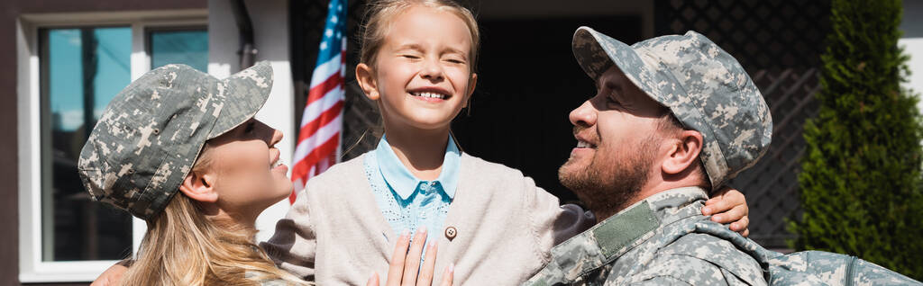 Gelukkige vader en moeder in militaire uniformen tillen dochter terwijl staan in de buurt van huis met de Amerikaanse vlag, banner - Foto, afbeelding