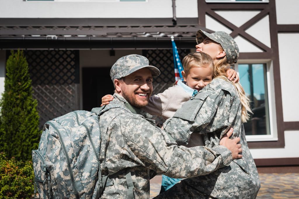 Fille souriante étreignant père et mère en uniformes militaires près de la maison avec drapeau américain - Photo, image