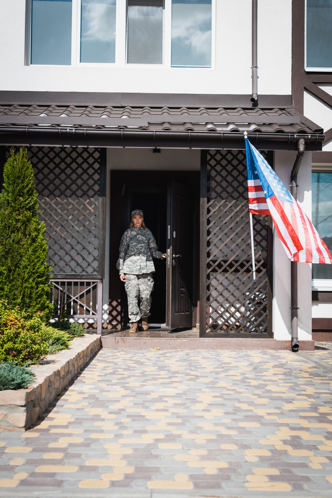 Katonai szolgálólány áll az ajtóban, közel az amerikai zászló és bokrok - Fotó, kép