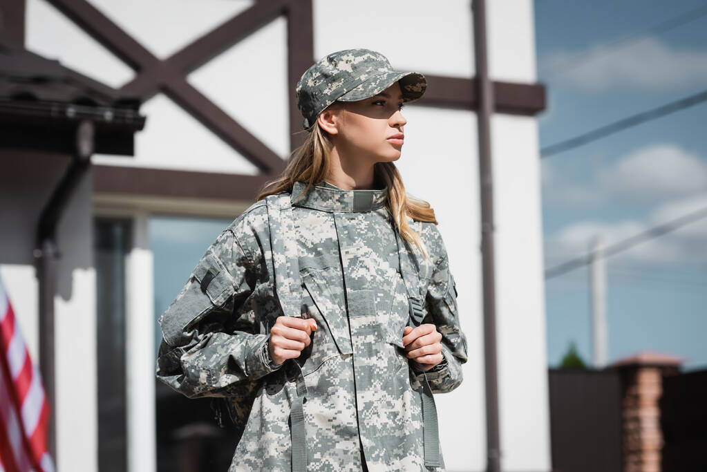 Femme de service militaire confiante avec sac à dos regardant loin avec maison floue sur fond - Photo, image