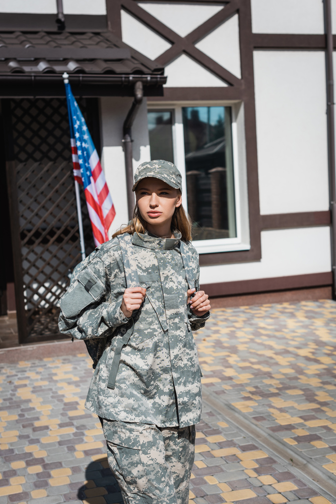 Ernste Soldatin verlässt Haus und schaut weg mit US-Flagge und Haus im Hintergrund - Foto, Bild