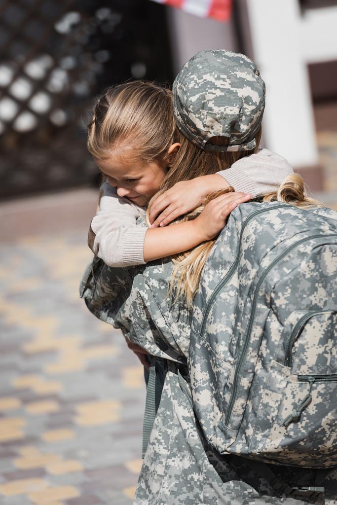 Sonriente hija abrazando a la madre en uniforme militar sobre fondo borroso - Foto, Imagen