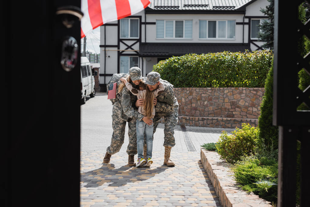Dochter knuffelen moeder en vader in militaire uniformen op straat in de buurt van huis op wazige voorgrond - Foto, afbeelding