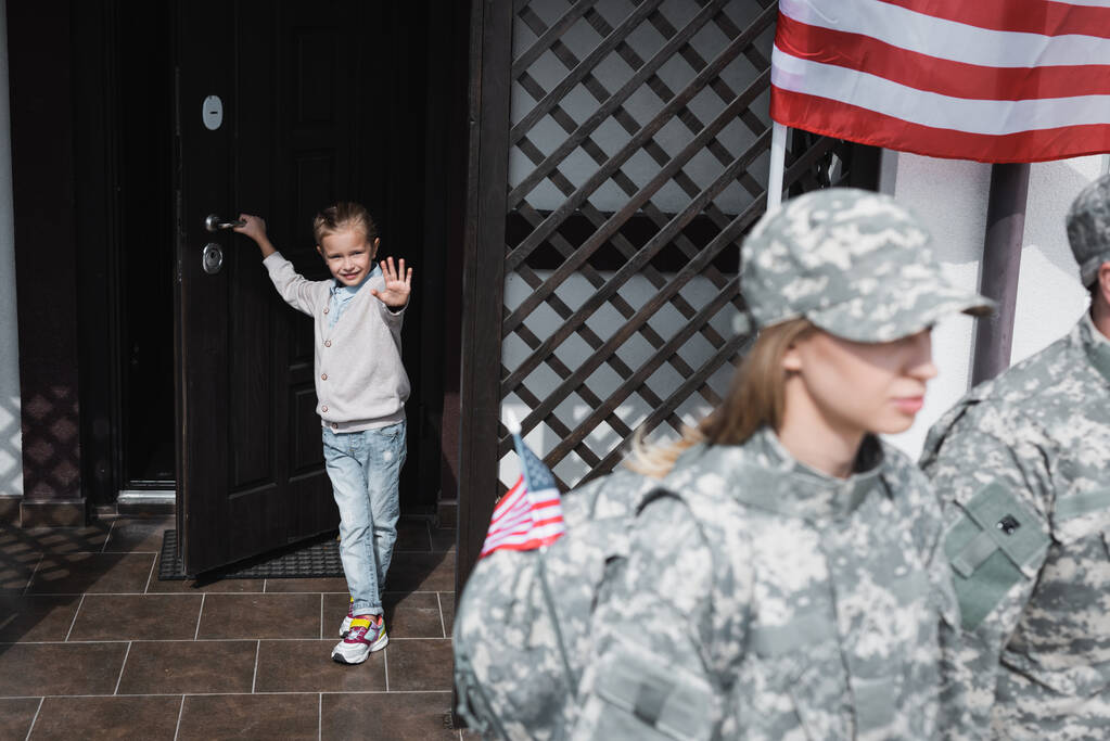 Sonriente hija con la mano ondeando de pie en la puerta cerca de la bandera americana con la madre borrosa y el padre en uniformes militares en primer plano - Foto, imagen