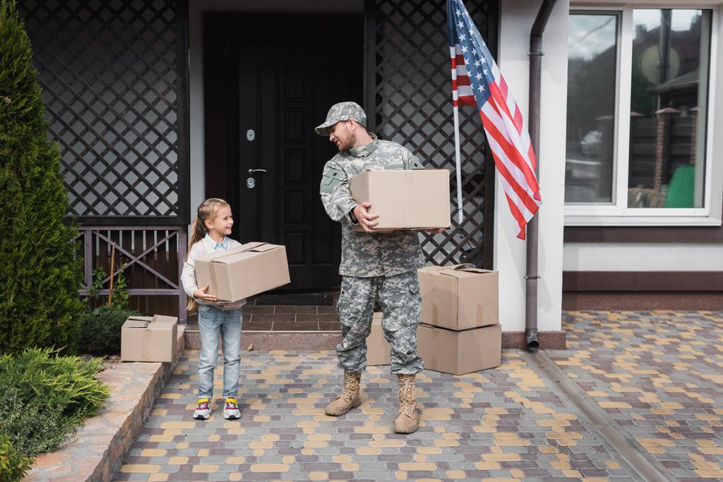 軍の制服を着た父とアメリカ国旗のある家の近くに段ボール箱を持っている娘 - 写真・画像