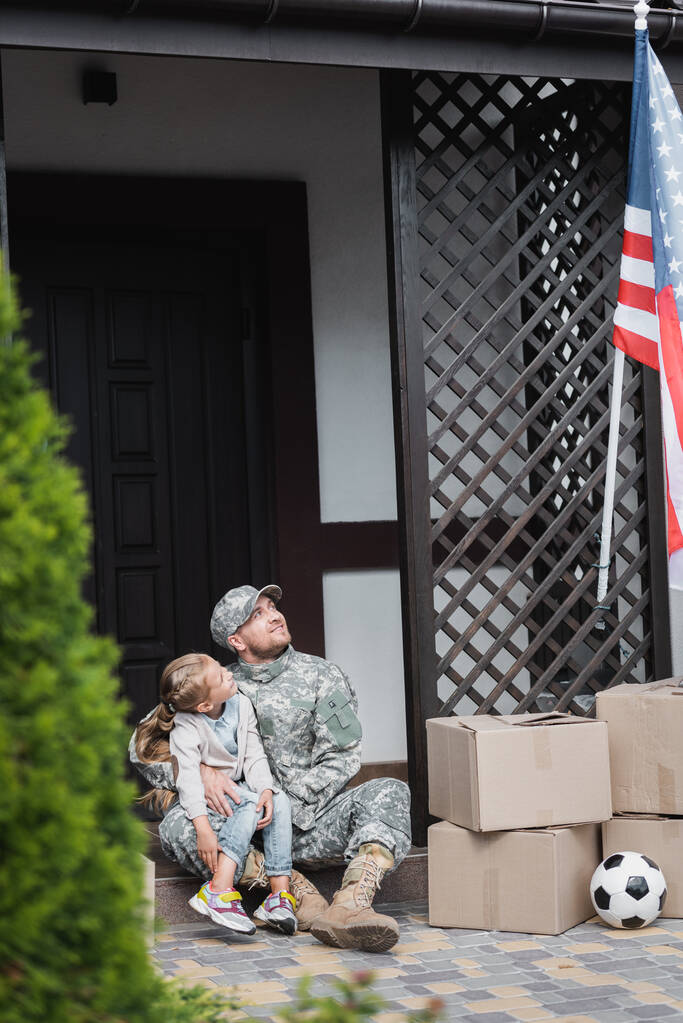 Apa katonai egyenruhában öleli a lányát, miközben ül a küszöbön, és nézi az amerikai zászlót  - Fotó, kép