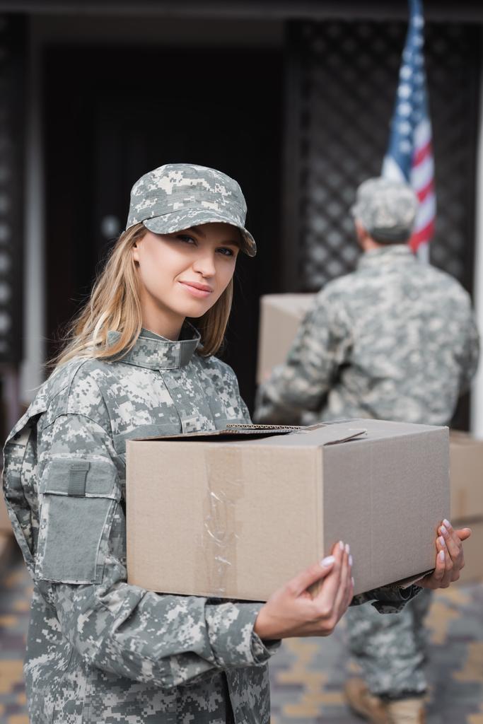 Mulher loira em camuflagem segurando caixa de papelão e olhando para a câmera com homem militar borrado no fundo - Foto, Imagem