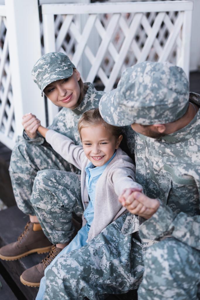 Счастливые военные отец и мать с дочерью сидят на пороге дома на размытом фоне - Фото, изображение