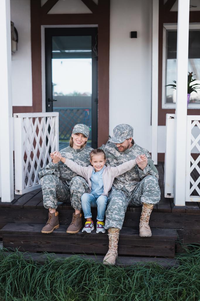Fille heureuse avec les mains tendues assis près de la mère et le père en uniformes militaires sur le seuil en bois - Photo, image