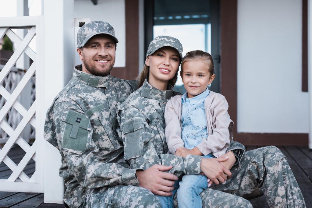 glimlachende militaire moeder en vader knuffelen met dochter, terwijl zitten op de drempel van het huis - Foto, afbeelding