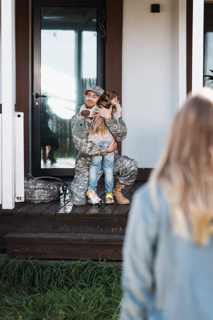 Lächelnder Militärmann umarmt Tochter, auf Knien sitzend, auf der Schwelle mit verschwommener Frau im Vordergrund - Foto, Bild