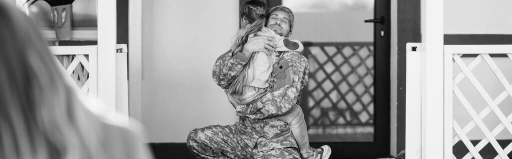 Padre en uniforme militar abrazando a la hija, mientras está sentado en la rodilla cerca de la puerta trasera con la mujer borrosa en primer plano, pancarta, monocromo - Foto, imagen