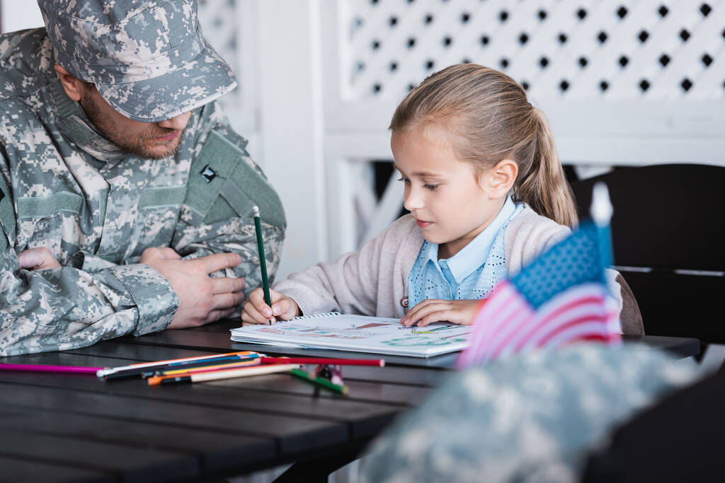 Батько у військовій формі сидить біля дочки малює барвистими олівцями вдома на розмитому передньому плані
 - Фото, зображення