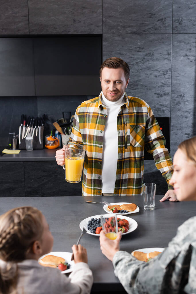 Uśmiechnięty mężczyzna z dzbankiem stojącym przy stole z naleśnikami i owocami z rozmazaną kobietą i dziewczyną na pierwszym planie - Zdjęcie, obraz