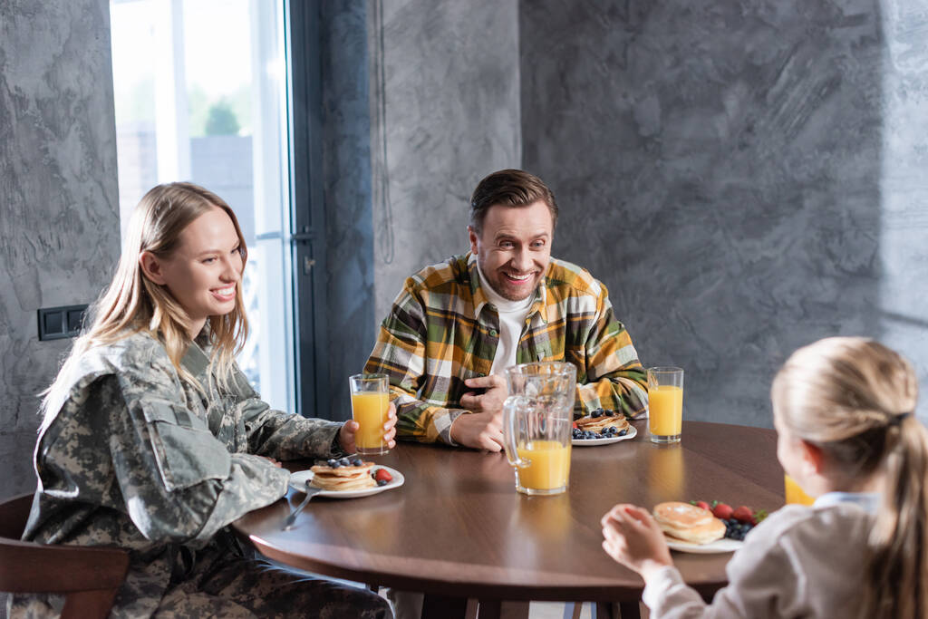 Szczęśliwa para jedząca śniadanie, śmiejąca się, siedząca przy stole w kuchni z zamazaną dziewczyną na pierwszym planie - Zdjęcie, obraz
