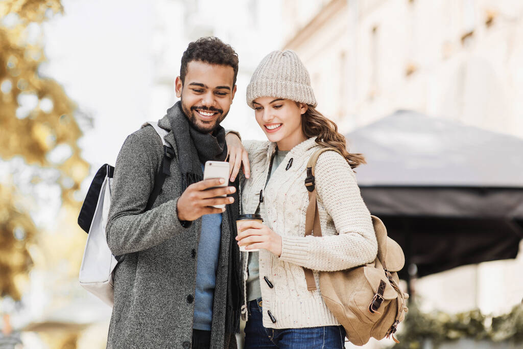 Piękna szczęśliwa para za pomocą smartfona. Młoda radosna uśmiechnięta kobieta i mężczyzna patrzący na telefon komórkowy w mieście jesienią. Technologia, podróże, turystyka, koncepcja studentów - Zdjęcie, obraz