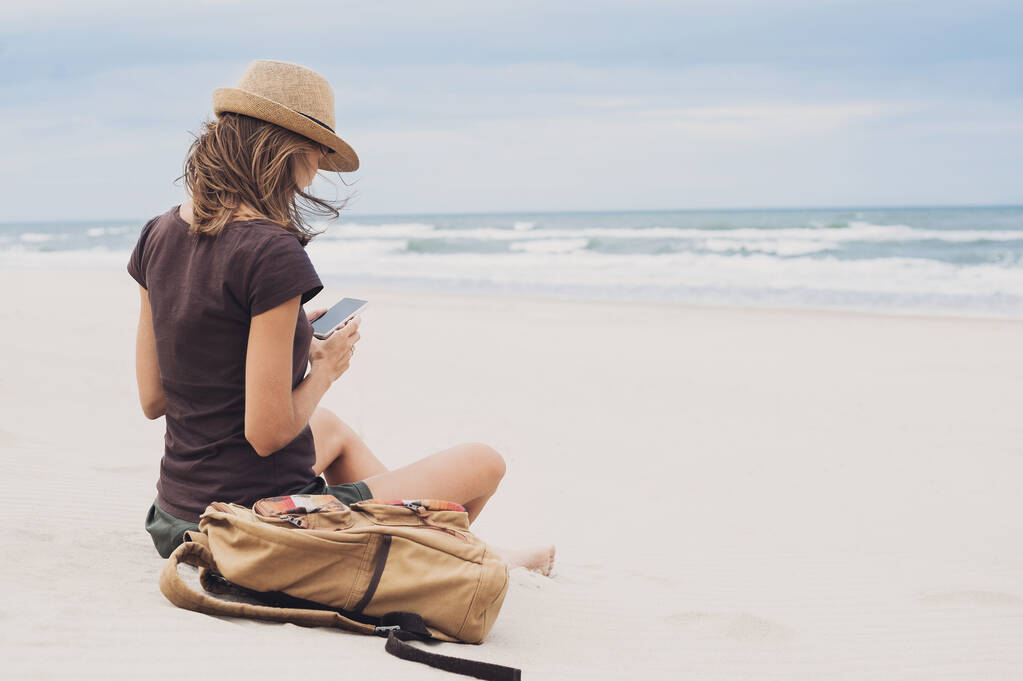Een jonge vrouw op het strand met een smartphone. Technologie en reisconcept. Mensen die mobiele apparaten gebruiken om vanuit afgelegen delen van de wereld verbonden te blijven - Foto, afbeelding