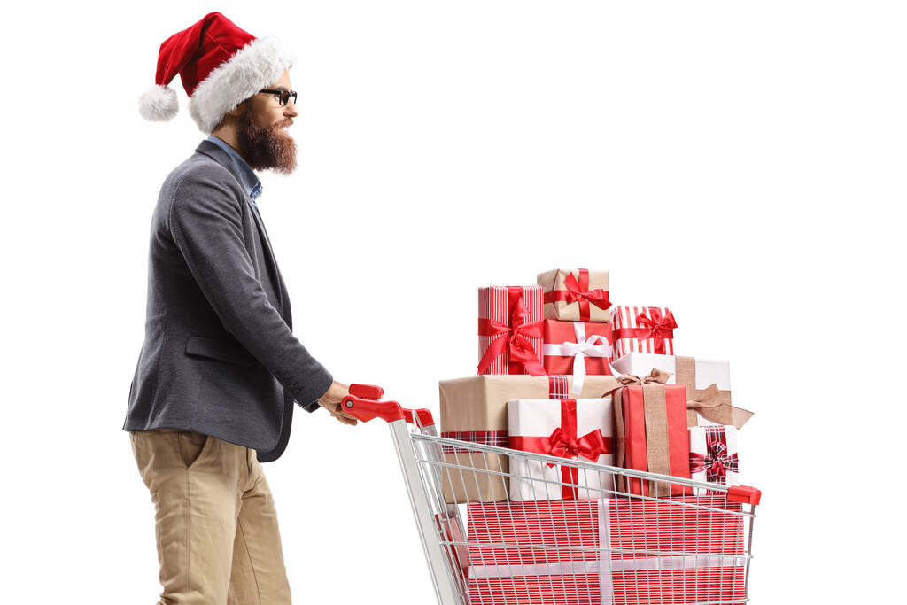 Homem com um chapéu de Papai Noel andando com um carrinho de compras cheio de presentes de Natal isolado no fundo branco - Foto, Imagem
