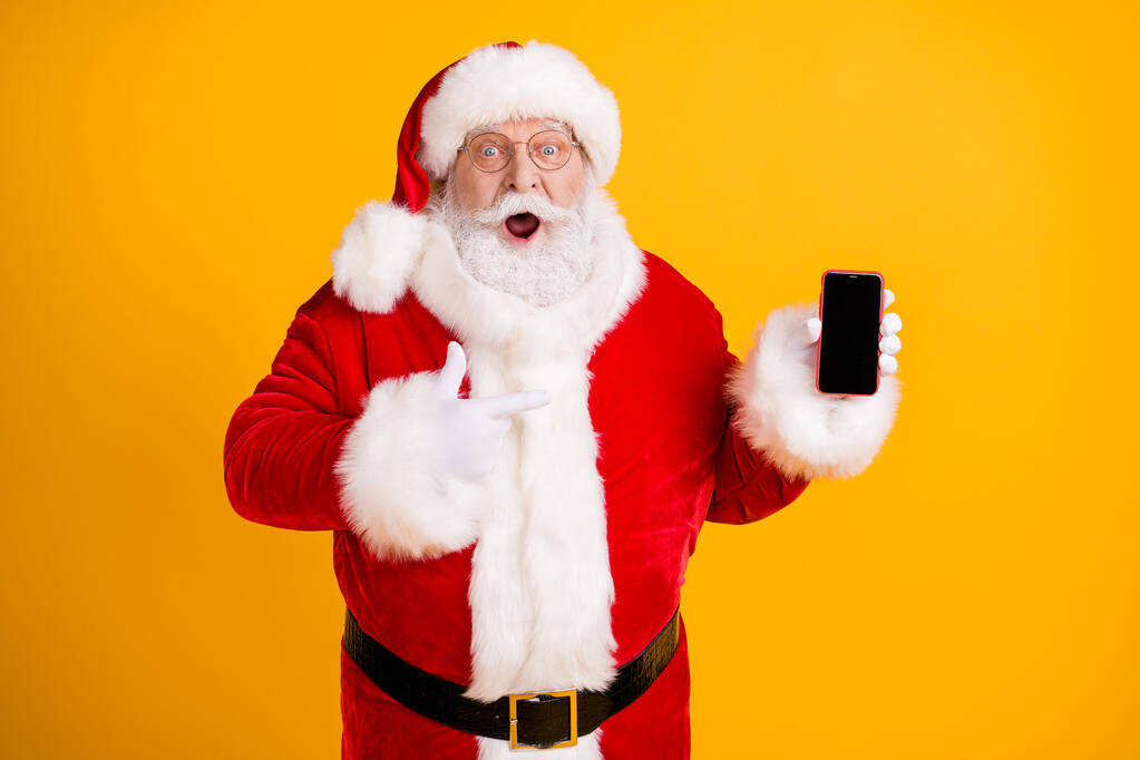Φωτογραφία λίπος άσπρα μαλλιά γκρι γενειάδα santa claus σημείο δάχτυλο smartphone εντυπωσιάστηκε καλύτερα Χριστούγεννα σεζόν ψώνια χριστουγεννιάτικο δώρο επιλογή φορούν κόκκινο καπέλο καπελάκι ρούχα απομονωμένο κίτρινο χρώμα φόντο - Φωτογραφία, εικόνα