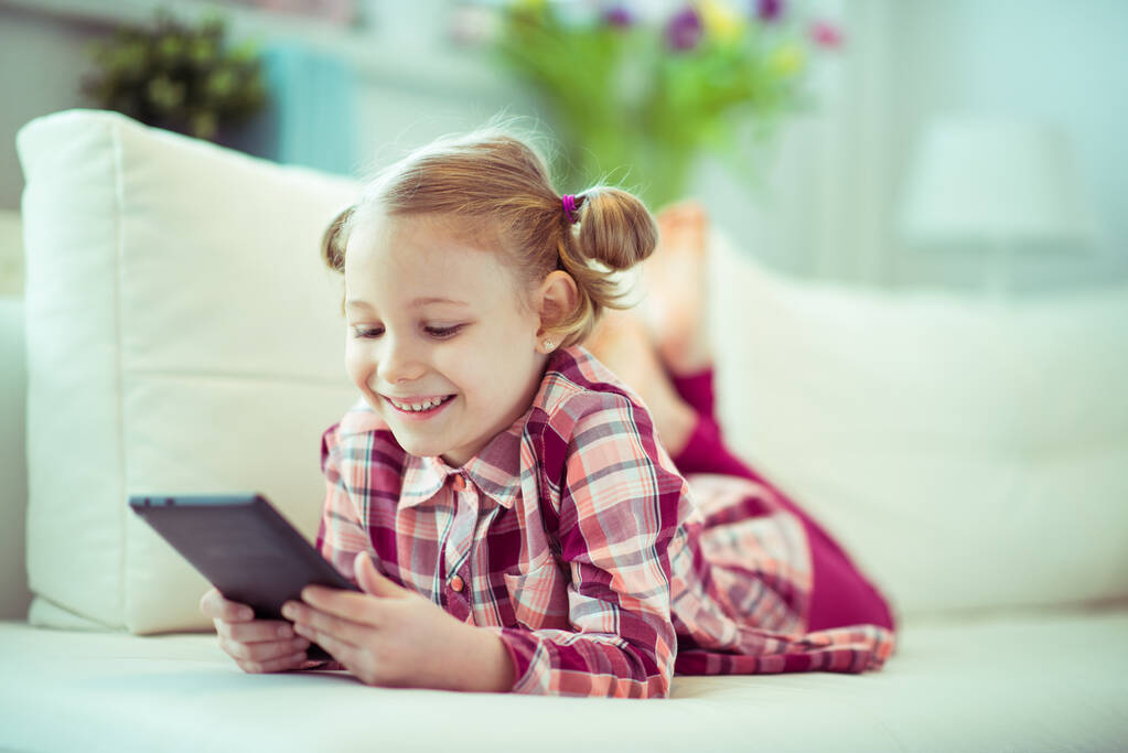 Retrato de una linda niña pequeña acostada en un sofá con una tableta en la educación en el hogar digital - Foto, Imagen