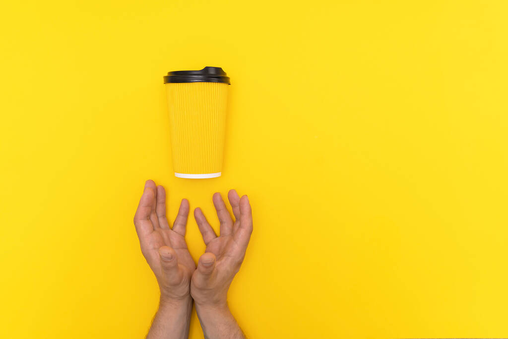 Dos manos masculinas caucásicas y taza de papel amarillo con tapa negra con café para llevar sobre fondo amarillo. - Foto, Imagen