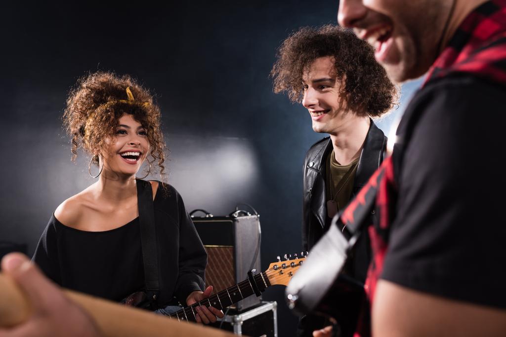 Musicista sorridente in piedi vicino a donna riccia con basso chitarra con chitarrista sfocato in primo piano - Foto, immagini
