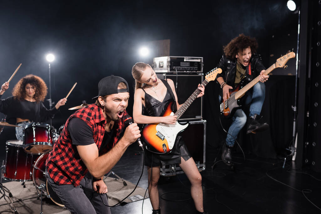 KYIV, UCRANIA - 25 de agosto de 2020: Vocalista gritando en micrófono inclinado hacia adelante cerca del guitarrista saltando durante el ensayo de la banda de rock con retroiluminación en negro - Foto, imagen