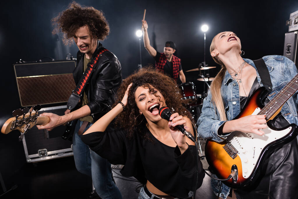 KYIV, UCRAINA - 25 AGOSTO 2020: cantante femminile di rock band con microfono che canta vicino a chitarristi con batterista sfocato sullo sfondo - Foto, immagini