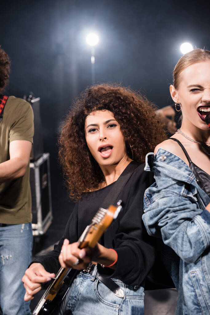 KYIV, UKRAINE - AUGUST 25, 2020: Curly vrouwelijke muzikant met open mond spelen elektrische gitaar staan in de buurt van vocalist met achtergrondverlichting op wazig achtergrond - Foto, afbeelding