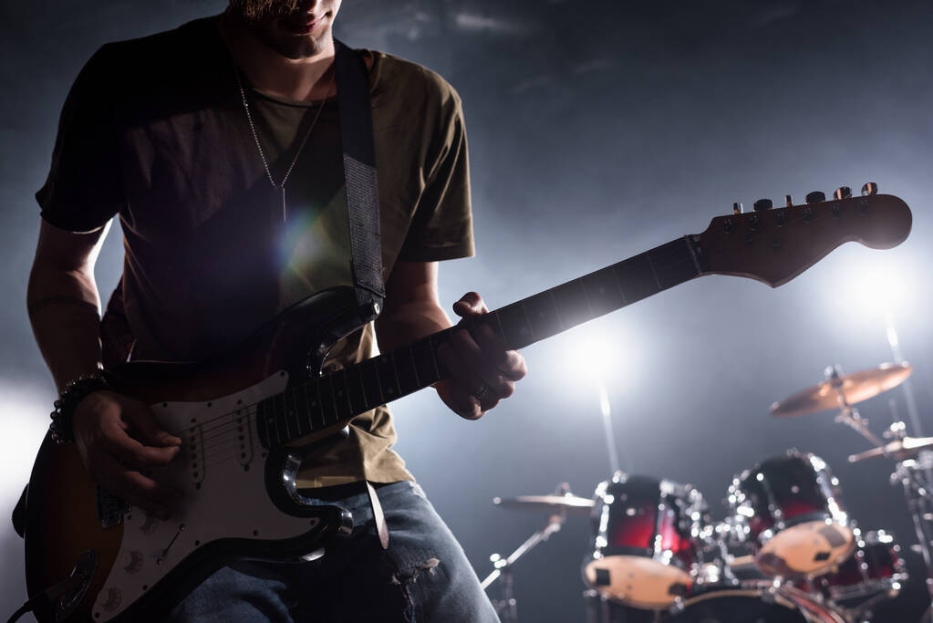 KYIV, UCRANIA - 25 de agosto de 2020: Vista recortada del músico de la banda de rock tocando la guitarra eléctrica con kit de batería borrosa en el fondo - Foto, imagen