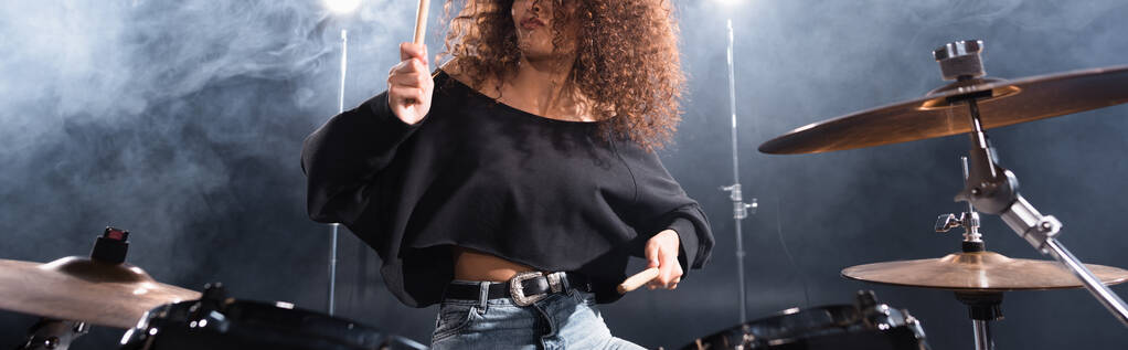 Gewassen weergave van krullende vrouw met drumsticks spelen op drum kit met rook op de achtergrond, banner - Foto, afbeelding