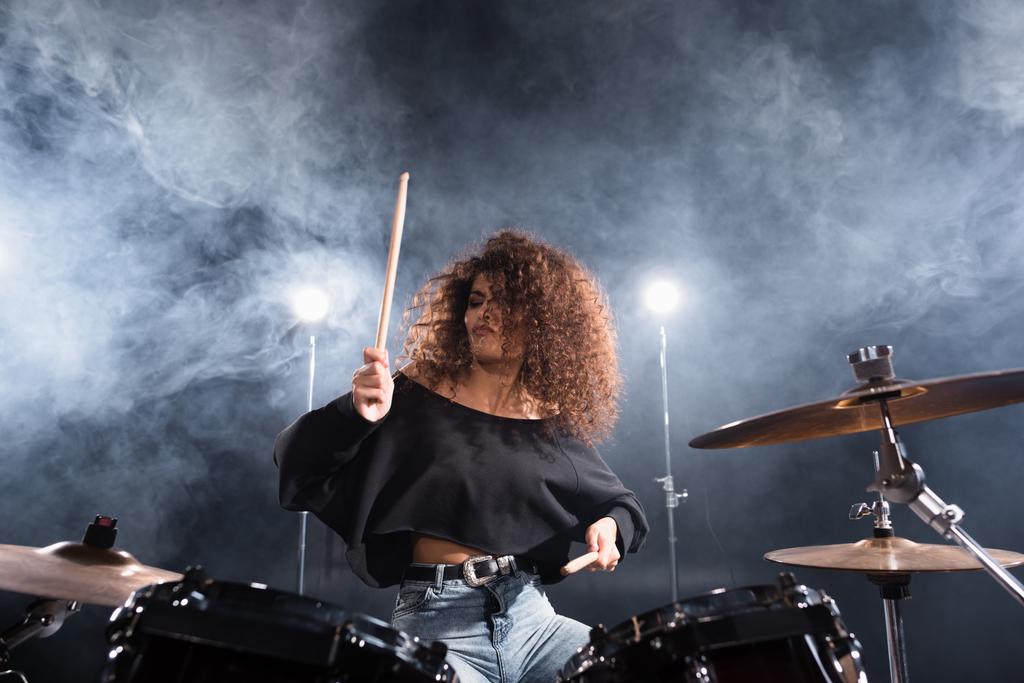 Кучерява жінка з барабанними паличками грає на барабанному наборі з димом і підсвічуванням на фоні
 - Фото, зображення