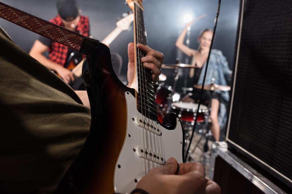 KYIV, UKRAINE - AUGUST 25, 2020: Close up view of man playing electric guitar near combo amplifiers with blurred drummer and guitarist on background - Photo, Image
