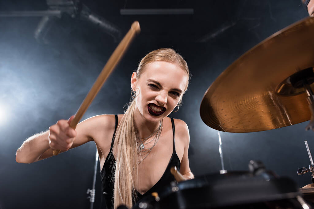 Blonde Frau mit Schlagstock zwinkert und spielt Schlagzeug auf verschwommenem Vordergrund - Foto, Bild