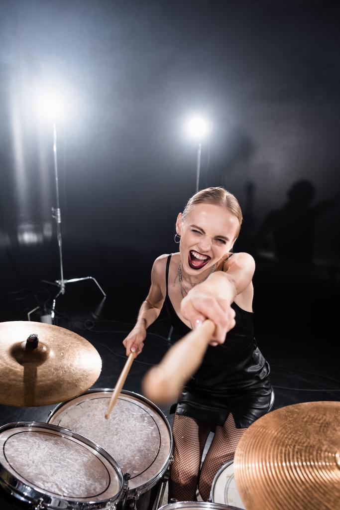 Hoge hoek uitzicht van opgewonden blond wijzen met drumstick tijdens het zitten op drum kit met achtergrondverlichting op wazig voorgrond - Foto, afbeelding