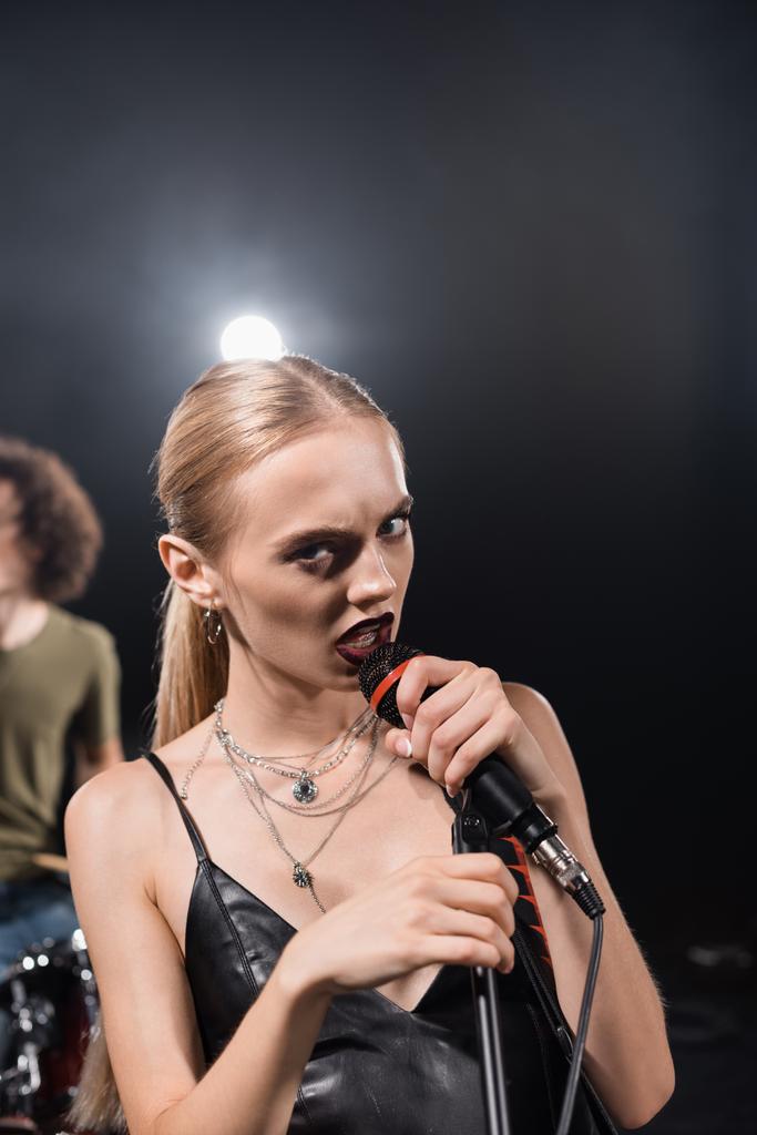 Θυμωμένη ξανθιά γυναίκα τραγουδώντας, ενώ αγγίζει ράφι μικρόφωνο με οπίσθιο φωτισμό και θολή ντράμερ στο παρασκήνιο - Φωτογραφία, εικόνα