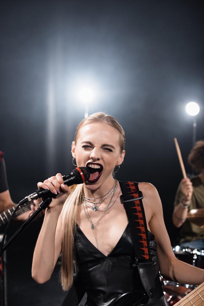 Блондинка с бас-гитарой кричит в микрофоне с подсветкой и размытыми музыкантами на заднем плане - Фото, изображение