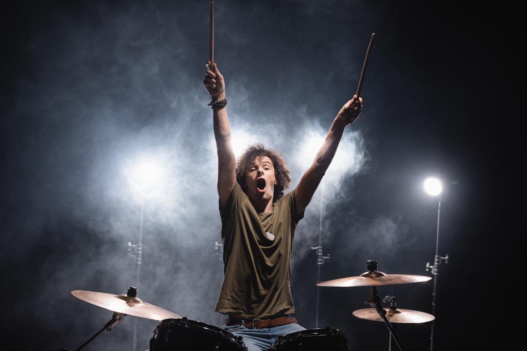 Кричащий музыкант с поднятыми руками сидит за барабанной установкой с дымом и подсветкой на заднем плане - Фото, изображение