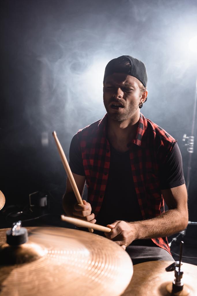Музыкант рок-группы держит в руках барабанные палочки и играет на барабанах с дымом на черном  - Фото, изображение
