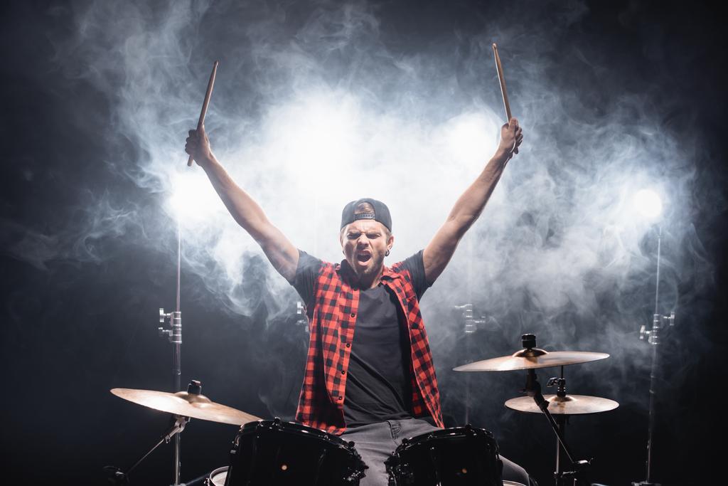 Кричащий барабанщик с поднятыми руками сидит за барабанной установкой с подсветкой и дымом на черном  - Фото, изображение