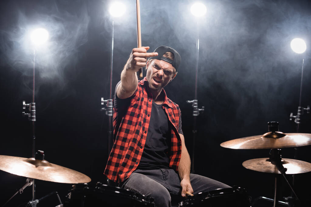 Злой музыкант показывает средний палец, держа в руках барабанные палочки и сидя за барабанной установкой с подсветкой на черном - Фото, изображение