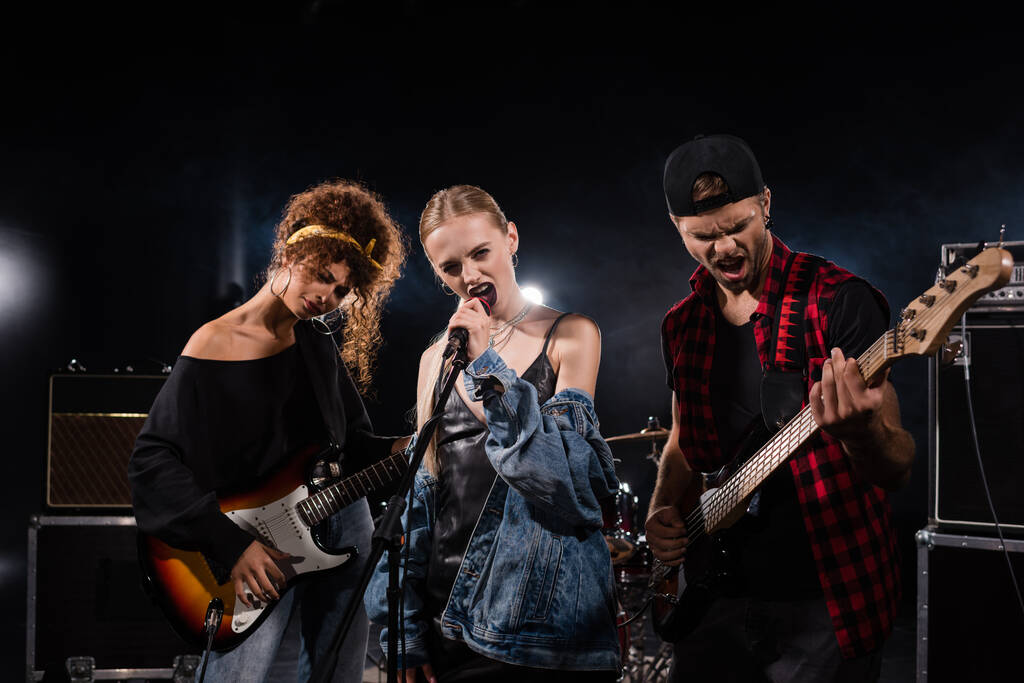 KYIV, UKRAINE - AUGUST 25, 2020: Blonde Вокаліст рок-гурту, який співає поруч з музикантами з бас-гітарами з підсвічуванням на чорному  - Фото, зображення