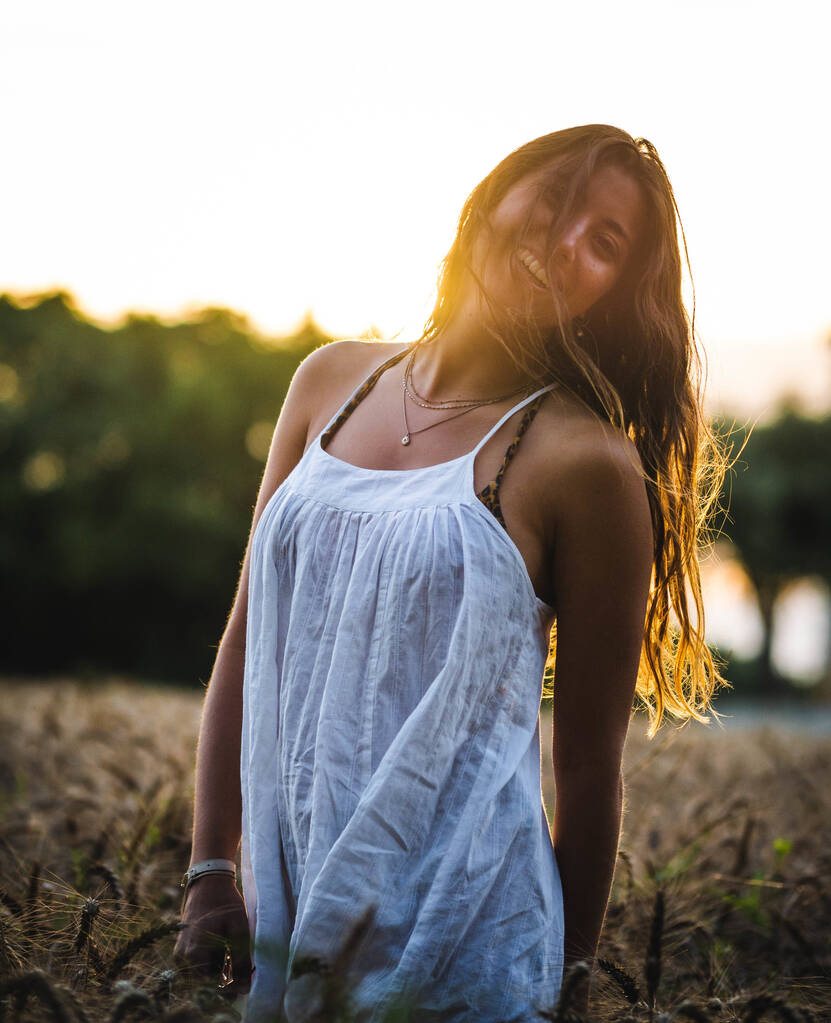 Μια κάθετη, ρηχή φωτογραφία μιας καυκάσιας γυναίκας με λευκό φόρεμα που ποζάρει σε χωράφι με σιτάρι το ηλιοβασίλεμα. - Φωτογραφία, εικόνα