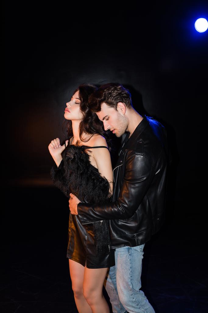 Appassionato uomo abbracciando fidanzata in abito toccare spalline con retroilluminato sul nero - Foto, immagini