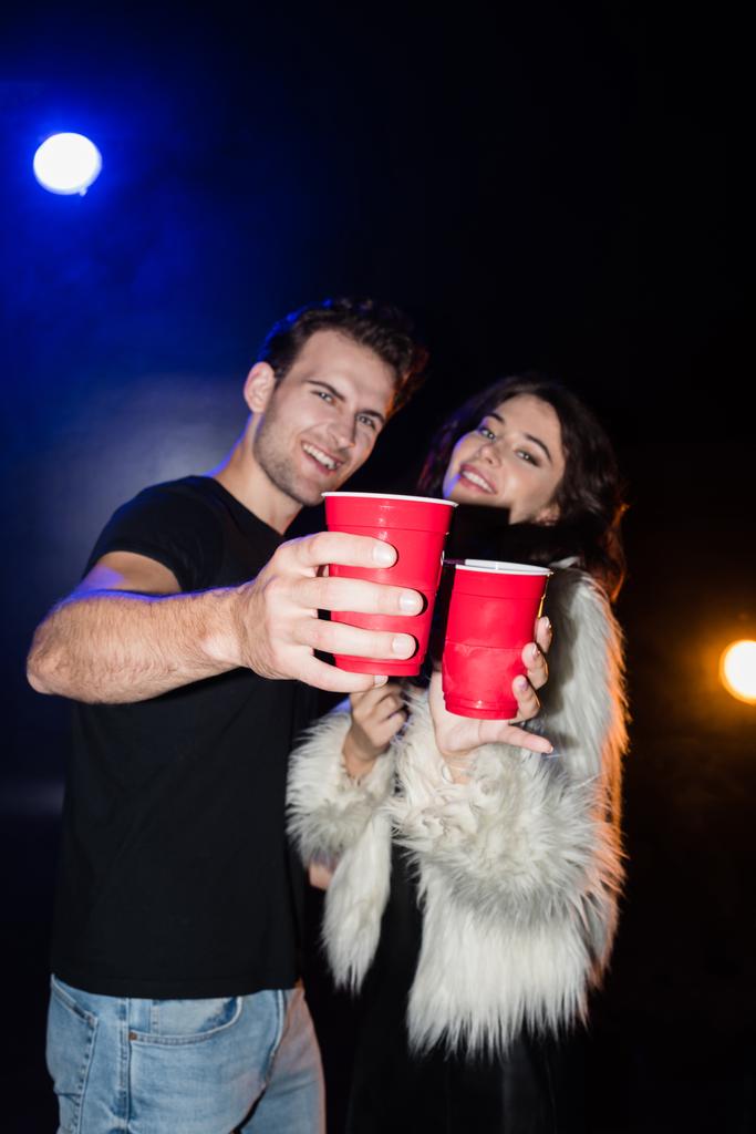 Счастливая пара, держащая красные пластиковые чашки и глядя на камеру с подсветкой на черном фоне, на размытом фоне - Фото, изображение